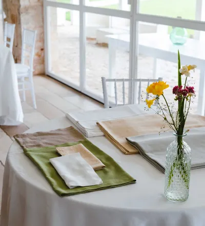 Tavolo rotondo con tovaglia bianca, tovaglioli verdi, beige, grigi e vaso di fiori gialli e fucsia in sala luminosa