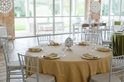 Tavolo rotondo con tovaglia color oro, tovaglioli piegati e sedie chiavarine bianche in una sala ricevimenti luminosa